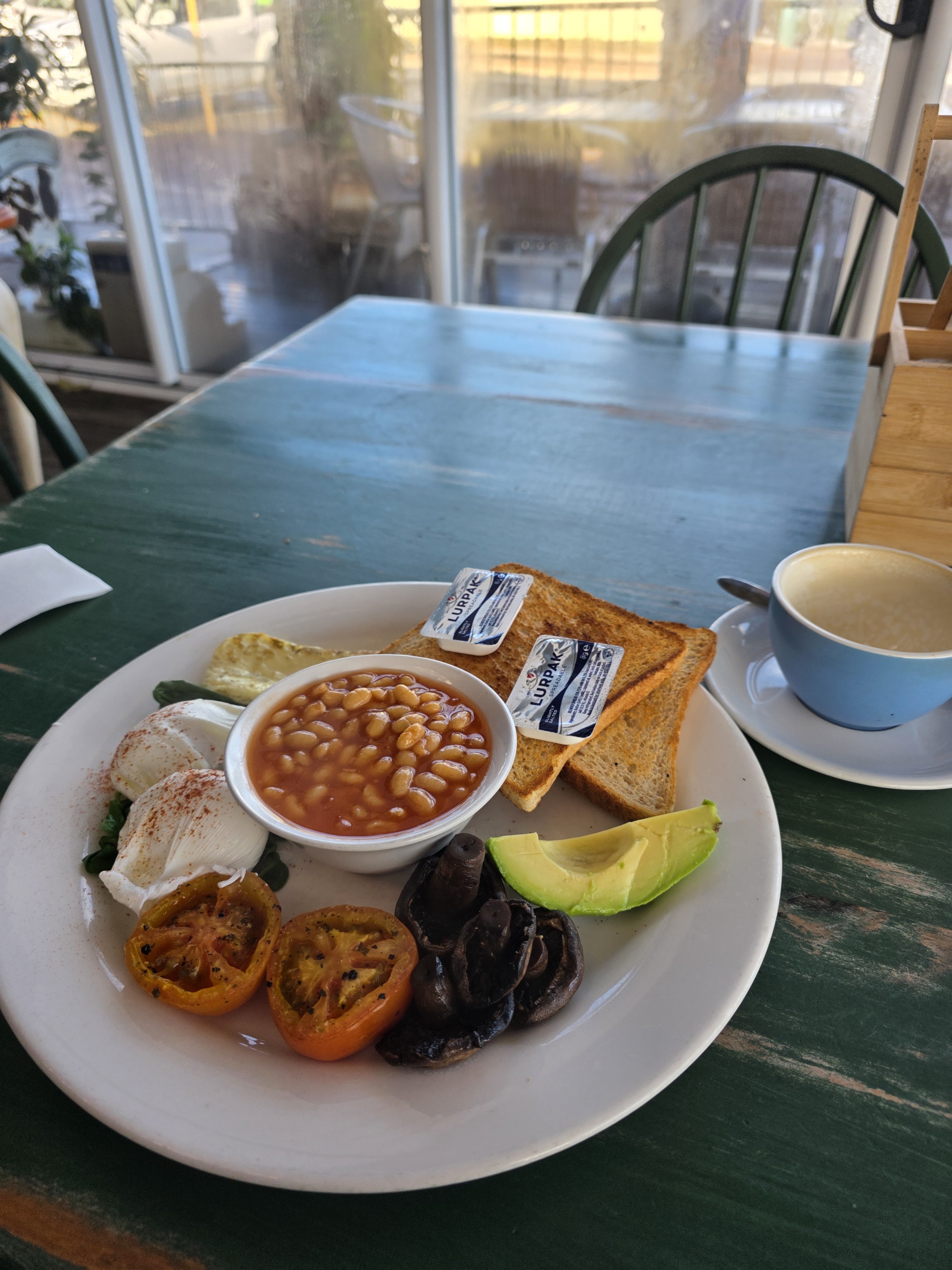 A breakfast plate featuring poached eggs, baked beans, grilled tomatoes, sautéed mushrooms, whole grain toast with butter packets, and a wedge of lime, set on a green wooden table.