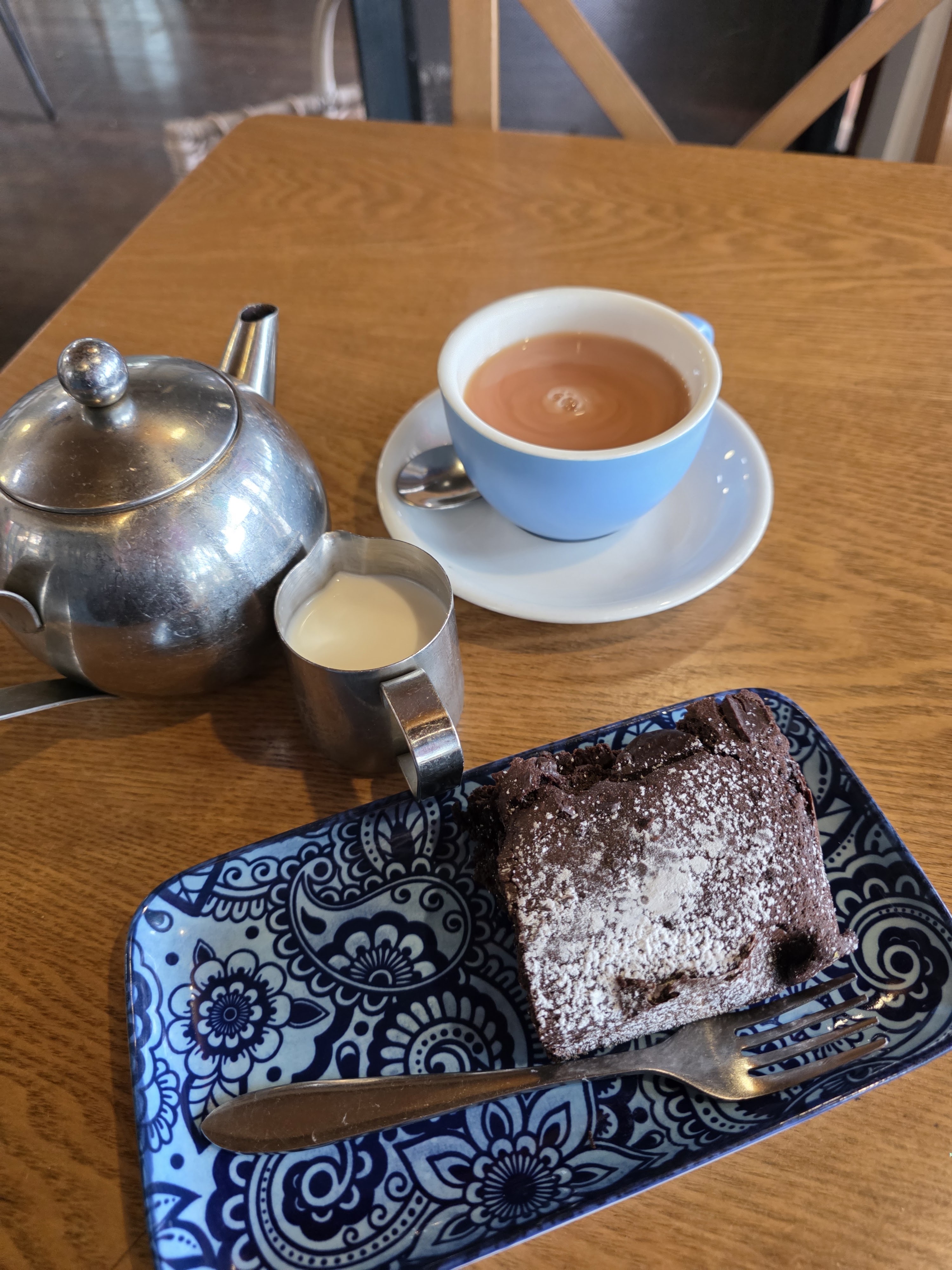 A silver teapot next to a blue cup of tea on a saucer, accompanied by a small pitcher of milk and a piece of chocolate cake on a decorative blue plate with a fork.