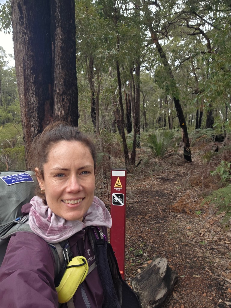 A woman with a backpack takes a selfie in a forested area, with tall trees and a hiking trail sign in the background.