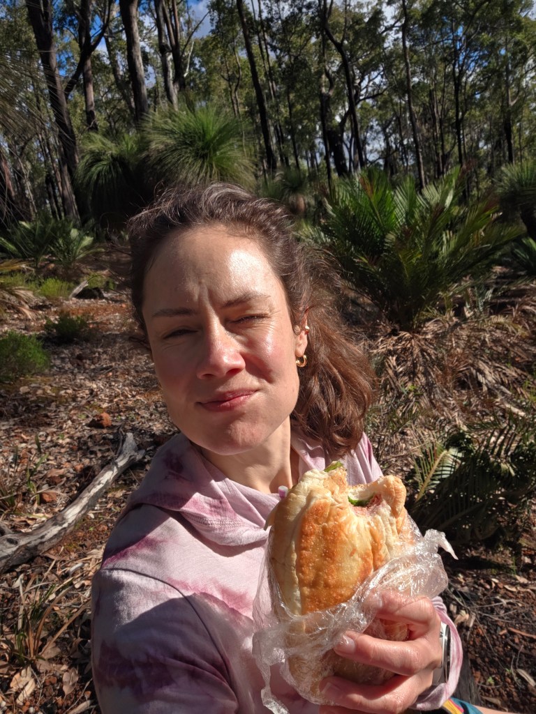 A person holding a sandwich, smiling in a forested area with trees and greenery in the background.