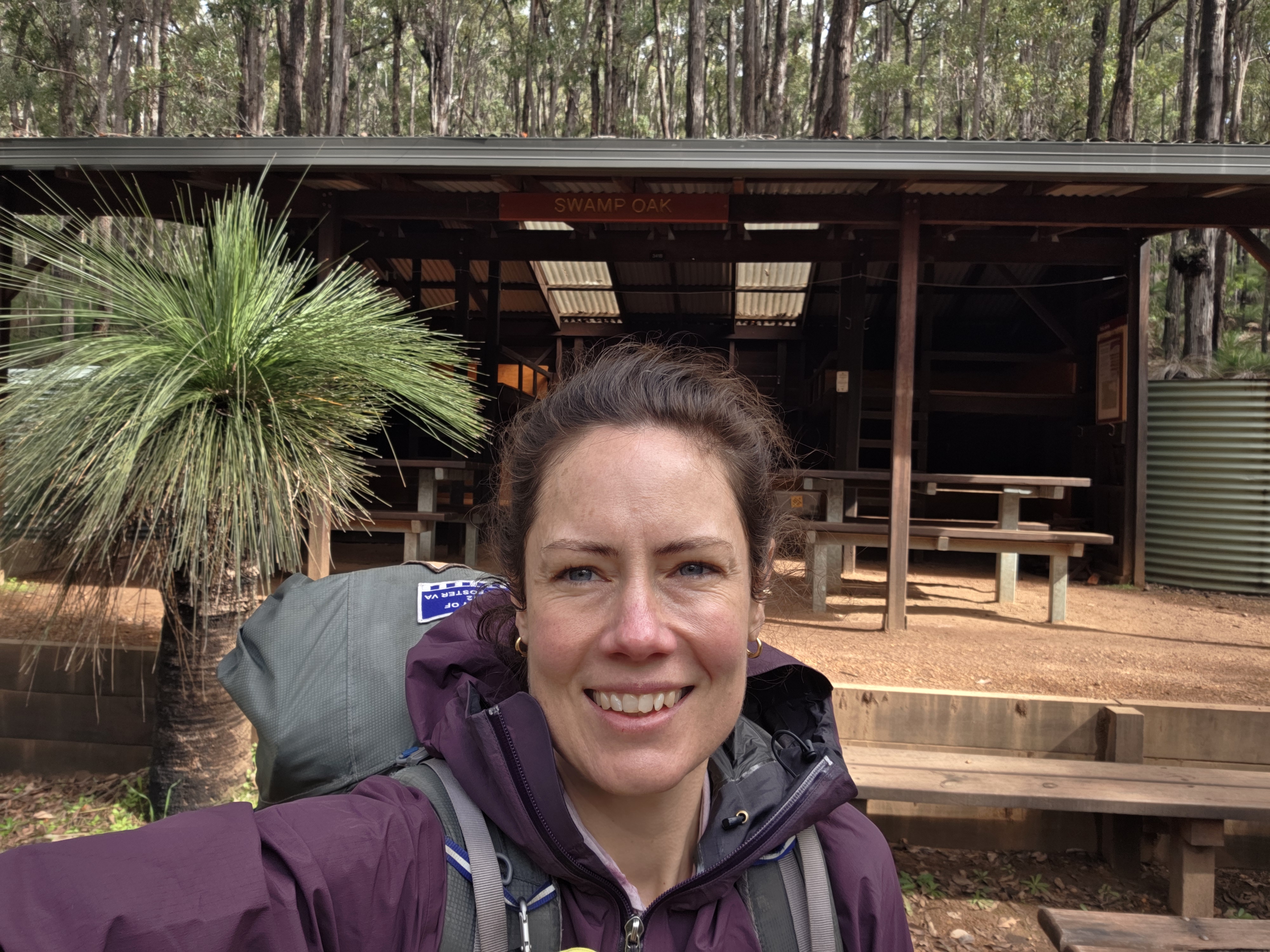 A person wearing a purple jacket and a backpack smiles in front of a rustic shelter surrounded by trees and vegetation.