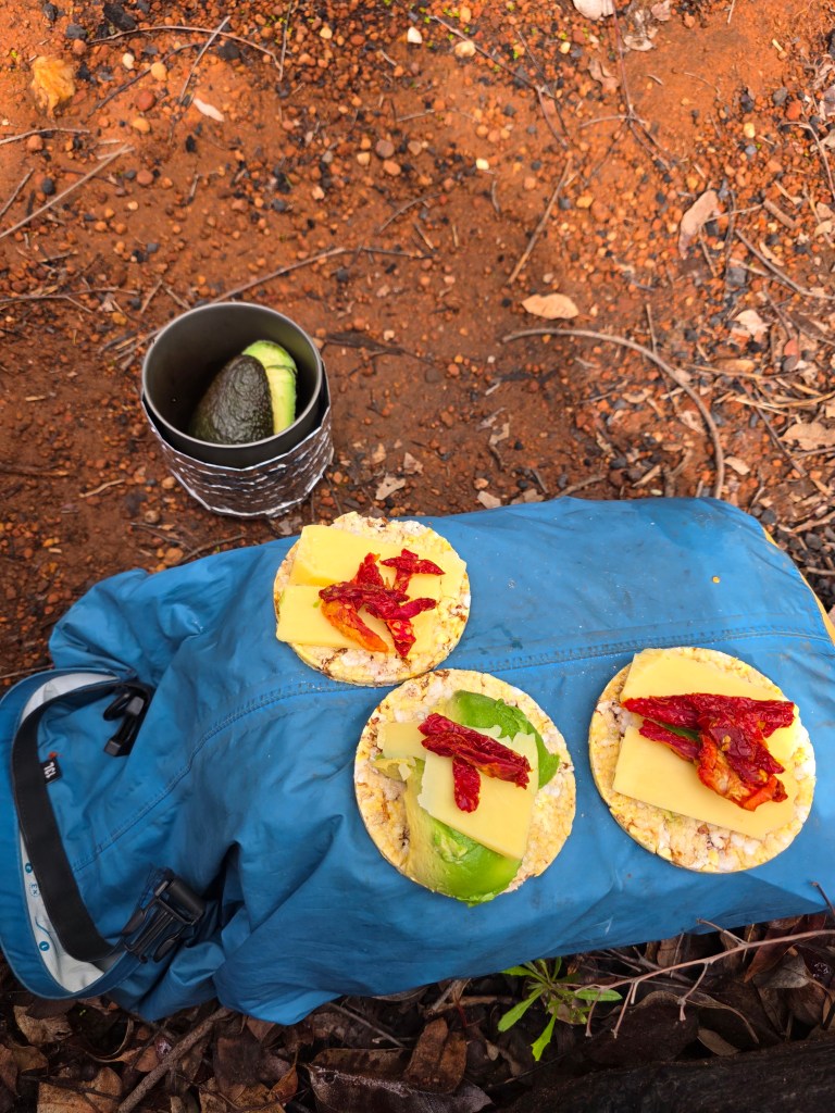Two open sandwiches with cheese, lettuce, and sun-dried tomatoes on a blue jacket, alongside a container holding an avocado, set against a dirt background.