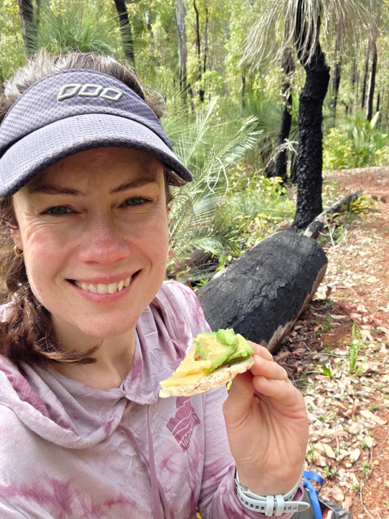 A woman with brown hair wears a cap and smiles while holding a slice of avocado and cheese on a cracker, sitting in a forested area with greenery and a charred tree in the background.