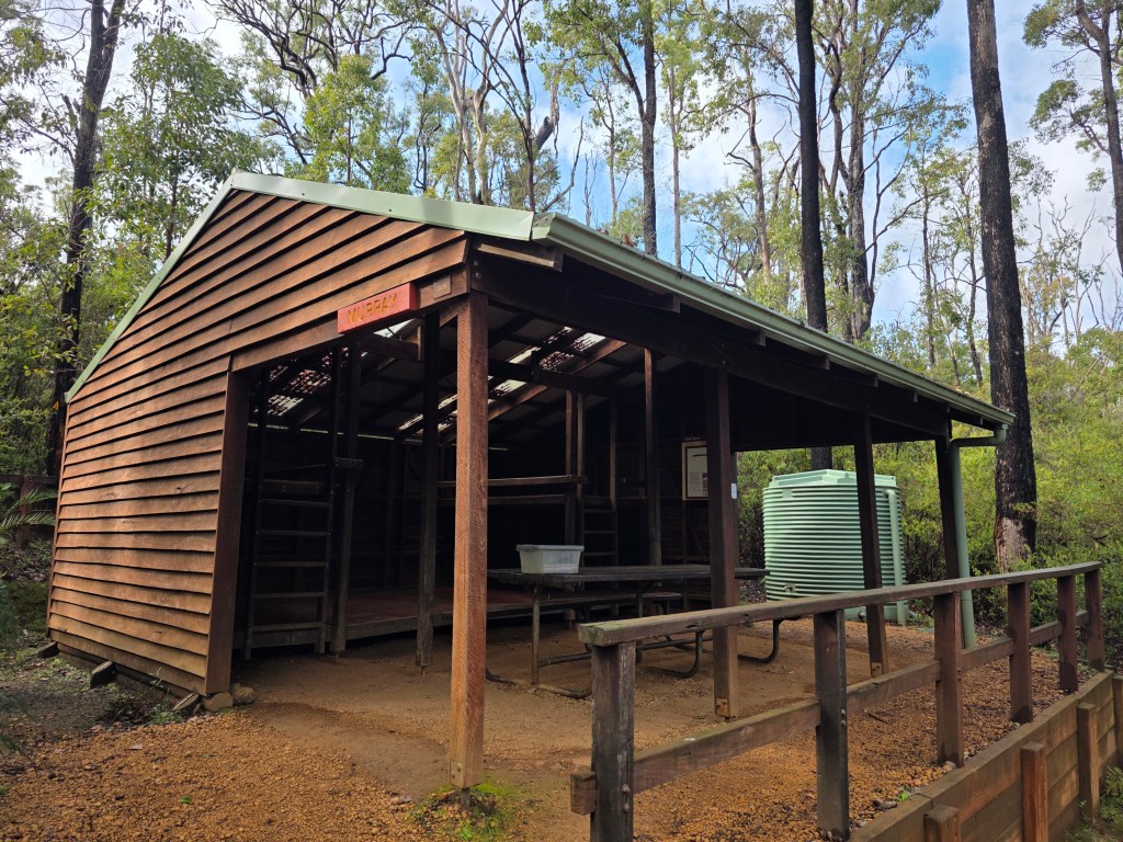 A rustic wooden cabin with a green roof, located in a forested area. The cabin features open sides, a sign labeled 'Murray', a bench outside, and a green water tank nearby.