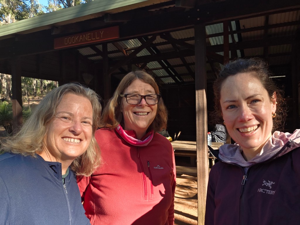 Three women smiling together in front of a wooden structure, with 'Dookanelly' sign above. They are outdoors, surrounded by trees.