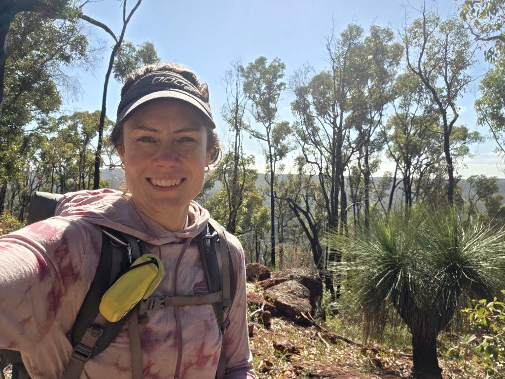 A hiker smiling for a selfie in a forested area, surrounded by trees and shrubs on a sunny day.