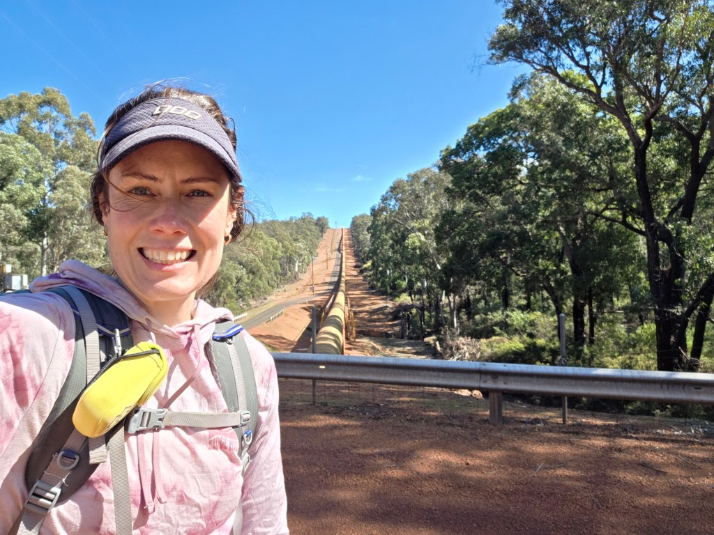 A woman smiling and posing for a selfie in front of a long pipeline running through a green and wooded area, with clear blue skies above.