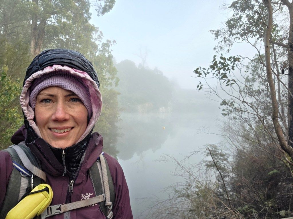 A woman in a purple jacket and beanie smiles at the camera while standing near a misty lake surrounded by trees.