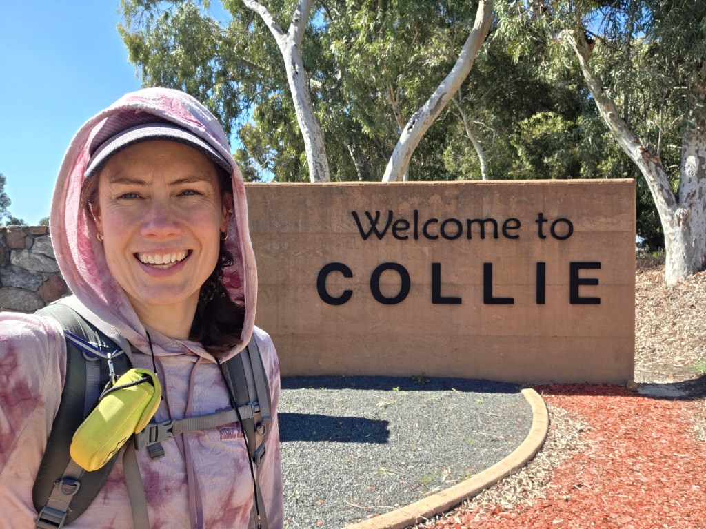 Person smiling in front of a 'Welcome to Collie' sign, surrounded by trees and a landscaped area.