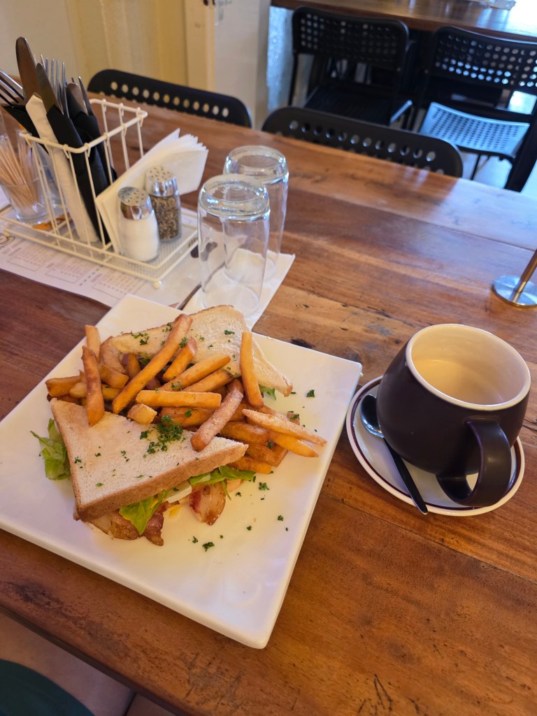 A plate featuring a sandwich with lettuce and bacon, topped with crispy fries, alongside a coffee cup on a saucer. The setting includes a wooden table with visible restaurant utensils and decor in the background.