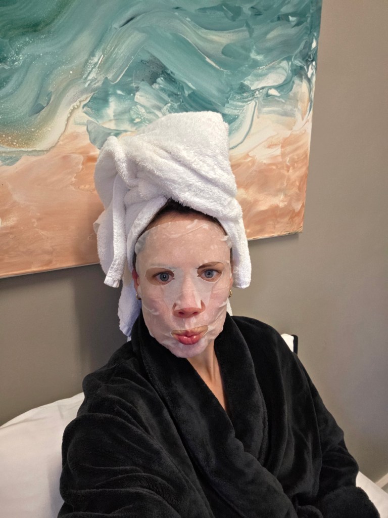 A person wearing a towel on their head and a facial mask, sitting on a bed, dressed in a black robe, with an abstract painting in the background.