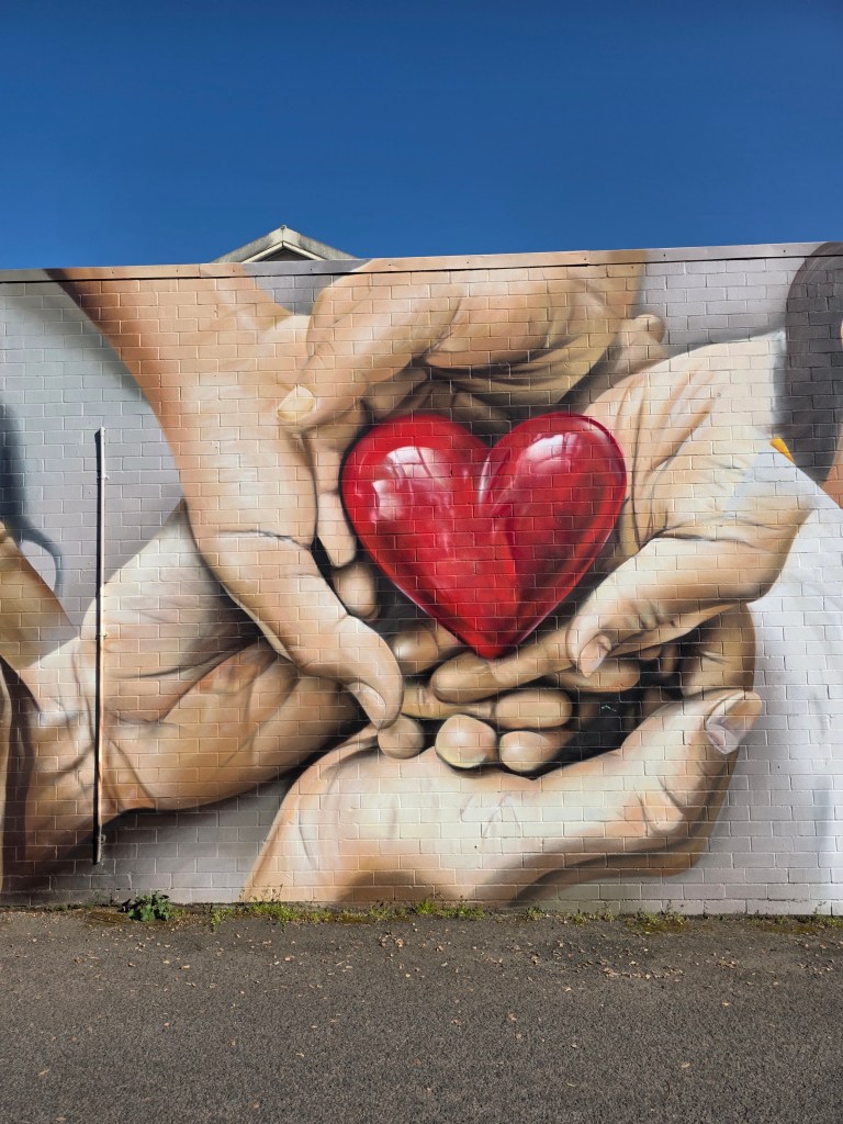 A mural depicting multiple hands of different sizes gently holding a large, shiny red heart.