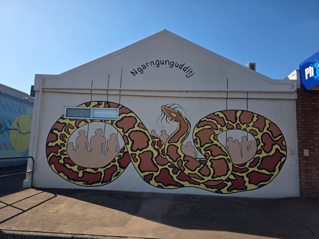 A large mural depicting a colorful snake intertwining around human figures, with the word 'Ngarngungudditj' displayed above on the wall.