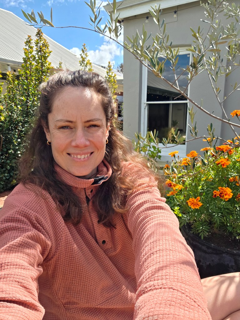 A woman in a light pink sweater takes a selfie in a garden filled with orange flowers and greenery, with a house in the background and blue skies above.