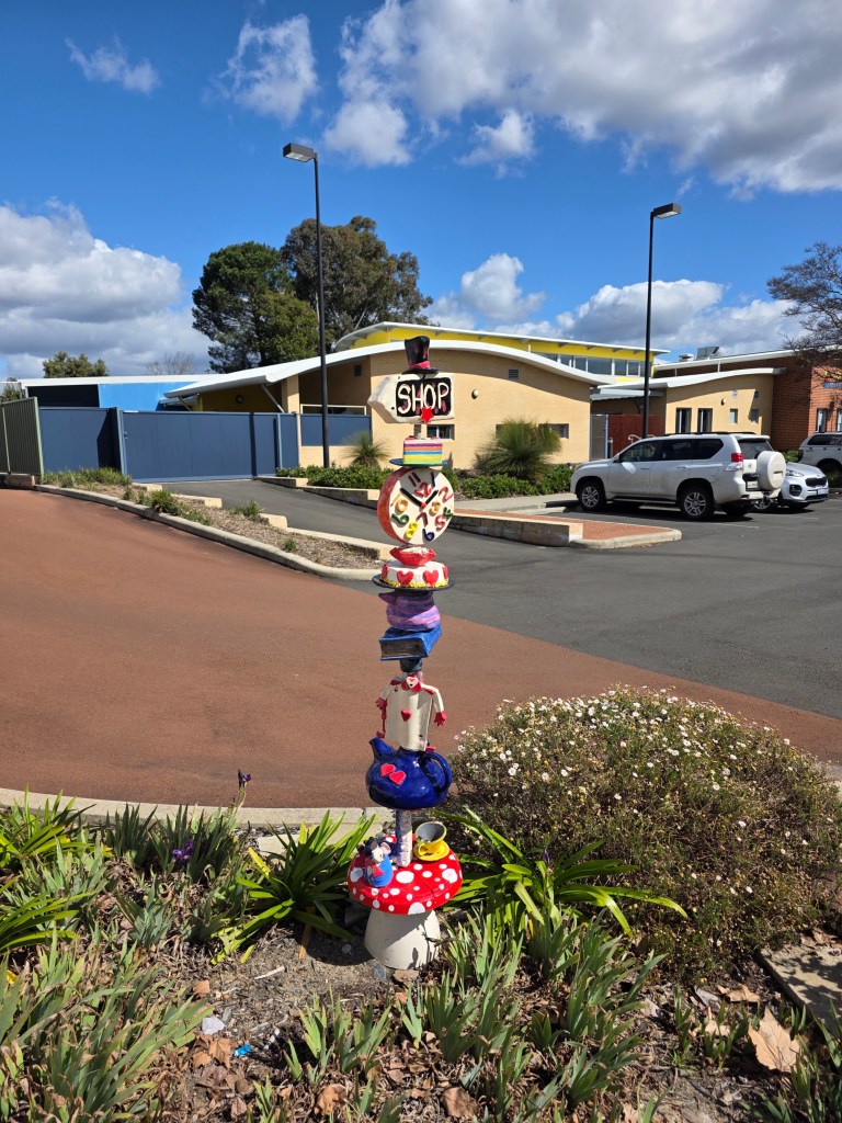 Colorful totem sculpture with painted figures and objects in a garden area, in front of a shop building under a blue sky with clouds.