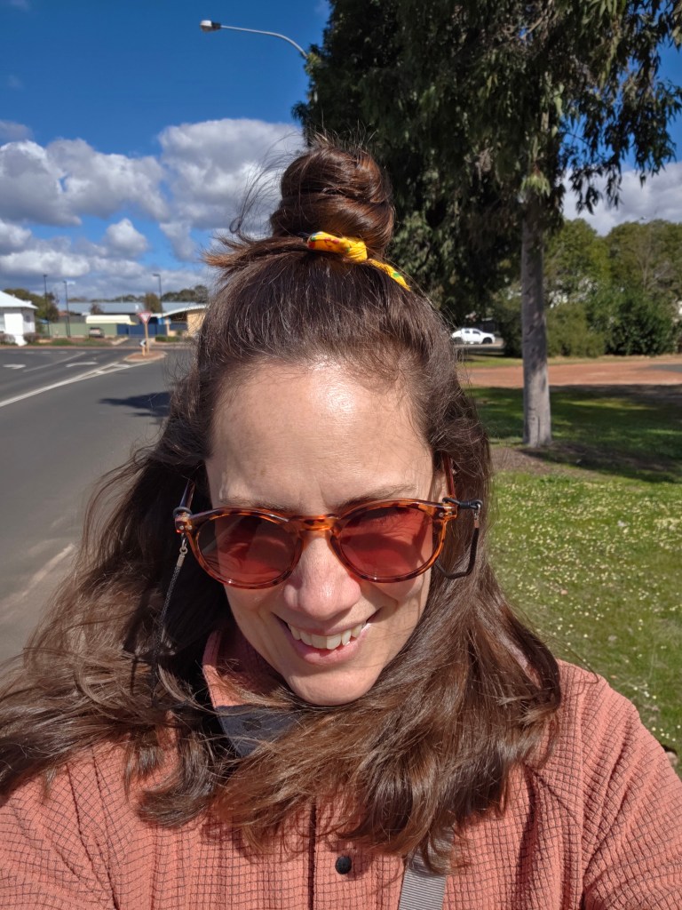 A woman with long hair styled in a bun, wearing sunglasses and a button-up shirt, smiles while standing on a road with a blue sky and scattered clouds in the background.